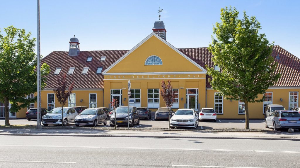 Hedelykke, gul murstensbygning med rødt tegltag og parkerede biler langs vejen.