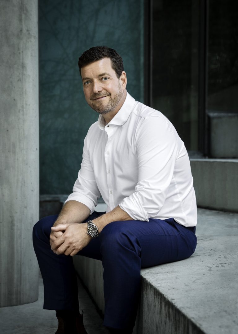 Lars Rosendahl i hvid skjorte siddende på en betonkant foran en mørk glasfacade.