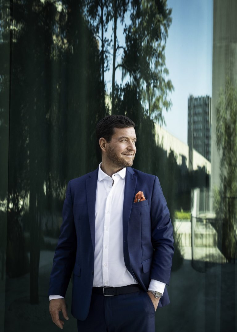 Lars Rosendahl i mørkeblåt jakkesæt og hvid skjorte foran en glasfacade, der spejler træer og himmel.